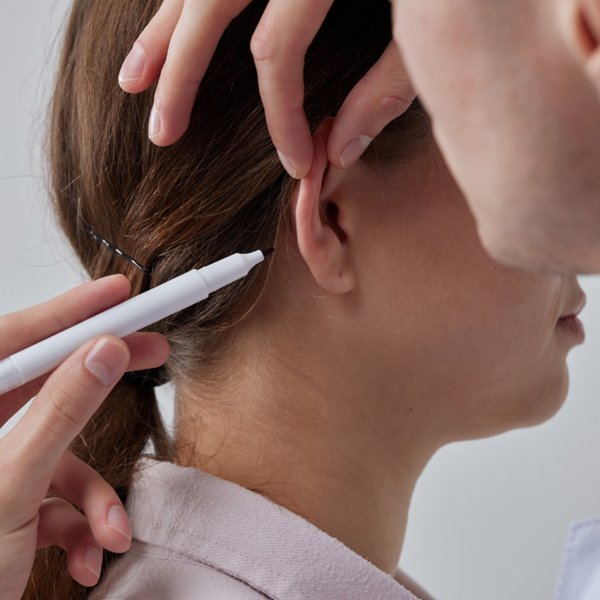A practitioner lightly marks behind a woman’s ear while holding it in place.