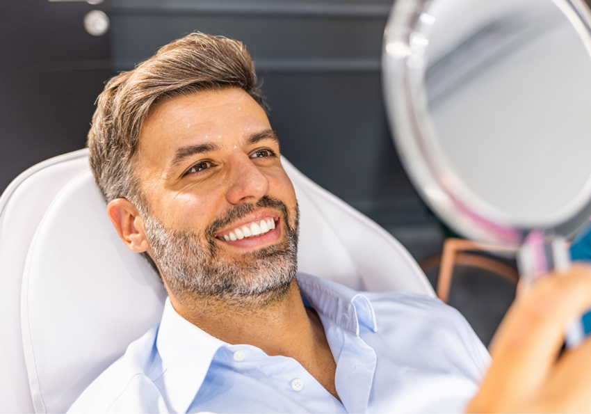 A man smiles while looking at his reflection in a handheld mirror.