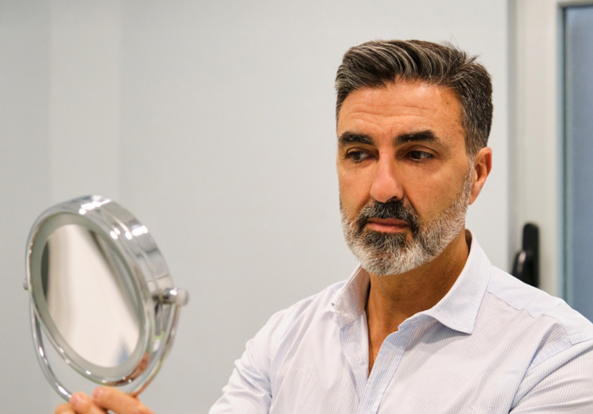 A man studies his reflection in a handheld mirror with a neutral expression.