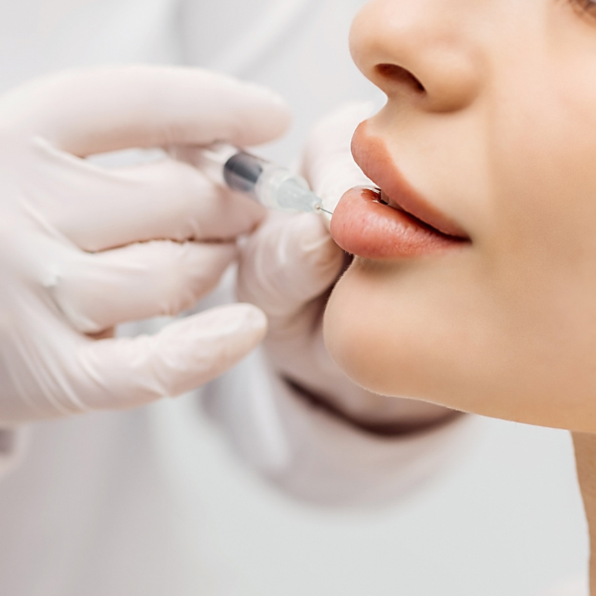 A close-up view shows a clinician injecting a woman’s lips with a syringe.