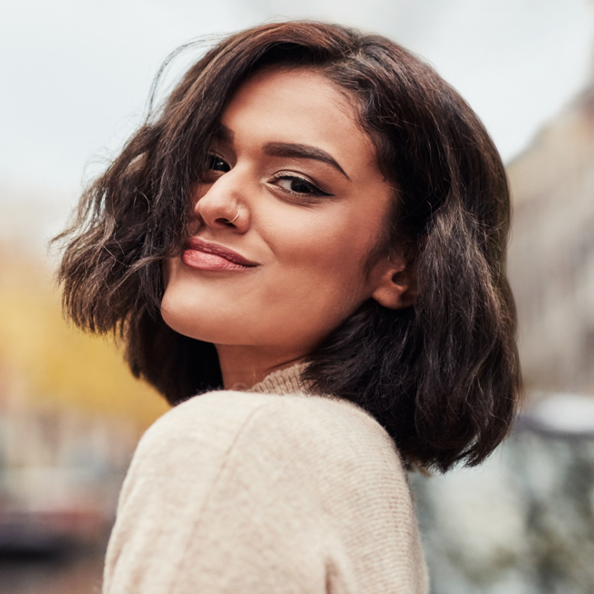 A woman with a short bob and nose ring smiles over her shoulder outdoors.