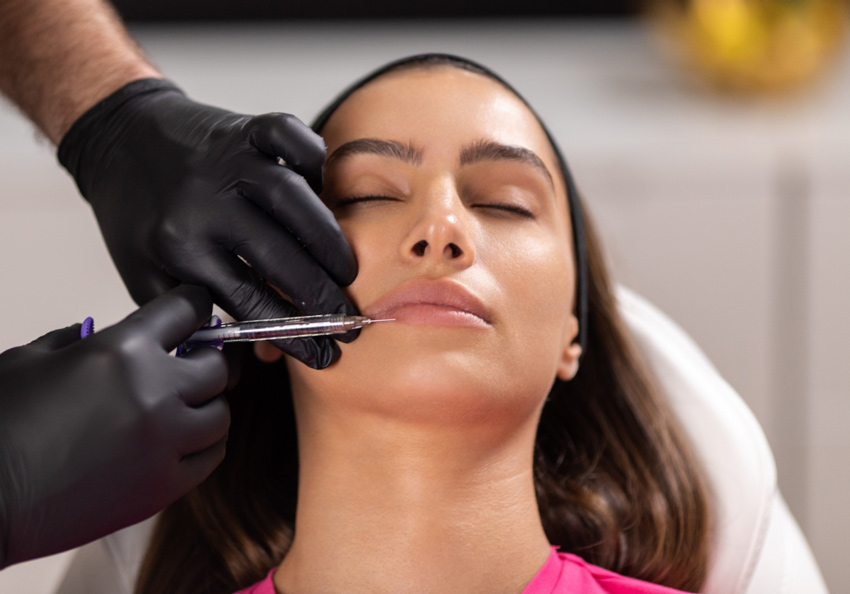 A woman lies relaxed with her eyes closed as a gloved practitioner works near her lips.