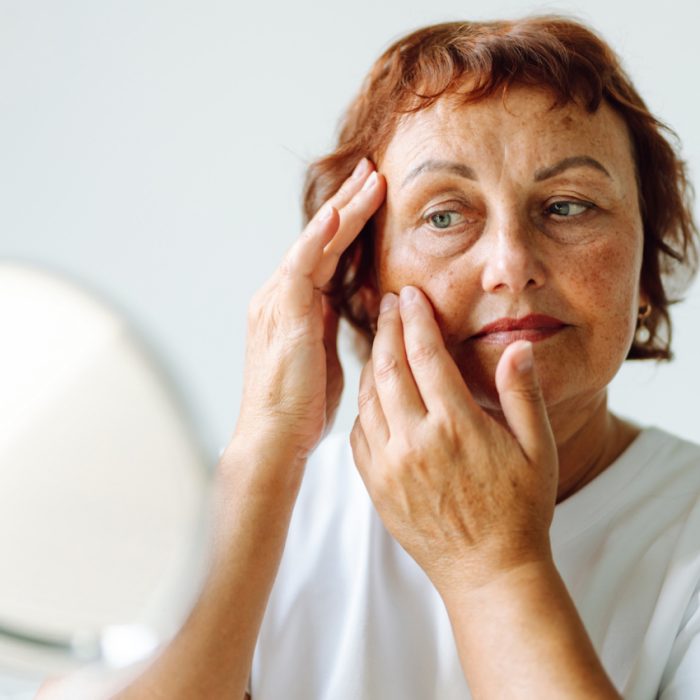 A woman with short auburn hair examines the skin on her face in a mirror.