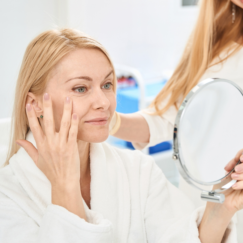 A woman in a white robe touches the skin near her eye while holding a handheld mirror.