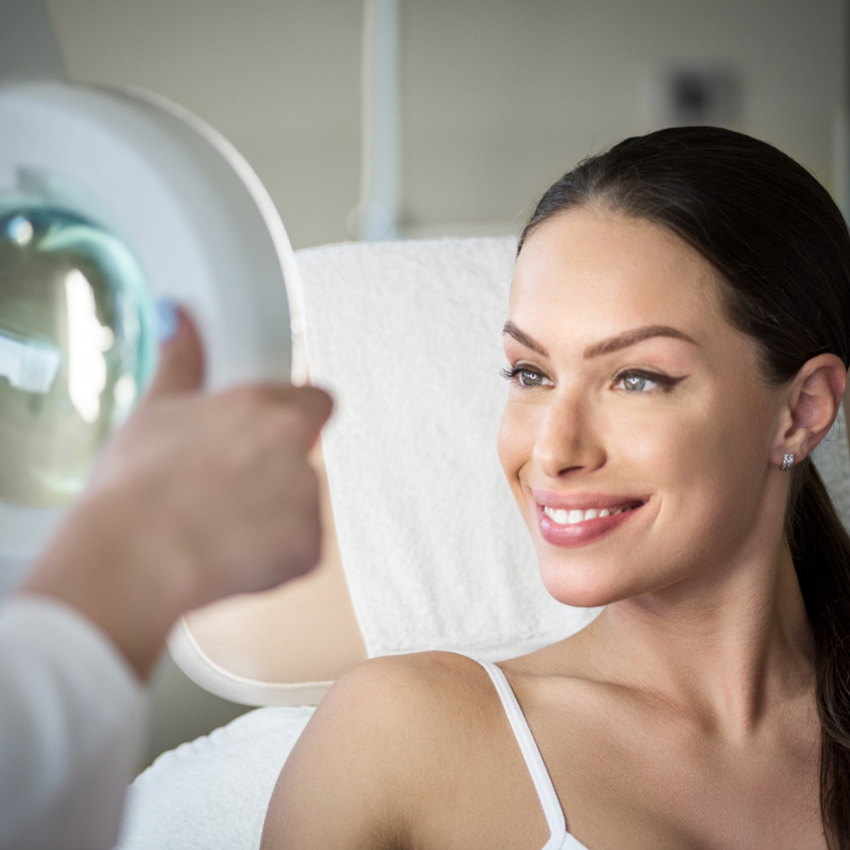 A woman smiles while looking into a handheld mirror during a skincare consultation.