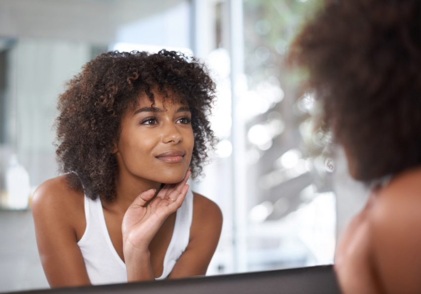 A woman looks at her reflection in a bathroom mirror while gently touching her chin.